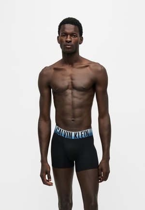 Modelo masculino en forma con piel oscura vistiendo calzoncillos bóxer negros de Calvin Klein con cinturilla azul con logotipo, de pie frente a un fondo blanco liso.