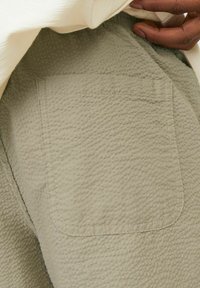 Gaiši zaļa teksturēta auduma drēbe ar aizdares kabatu ar šuvēm malās. Materiāls ir ar smalku, izvirzītu rakstu, kas piešķir vizuālu interesi.