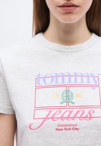 Νεαρό άτομο που φοράει ανοιχτό γκρι T-shirt με πολύχρωμο λογότυπο «tommy jeans» και κείμενο «Established New York City 1985» μπροστά.