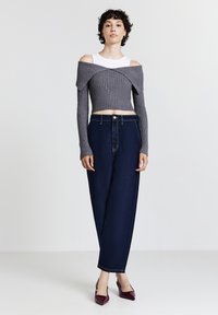 Frau trägt einen grauen, schulterfreien, gerippten Crop-Pullover über einem weißen Tanktop, dunkelblaue, hoch sitzende Jeans und kastanienbraune, spitze Absätze.