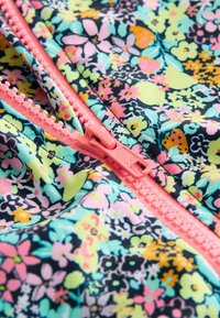 Nahaufnahme eines korallenrosa Reißverschlusses auf einem Stoff mit einem dichten floralen Muster in Pastellgelb, -rosa, -orange, -türkis und Schwarz.