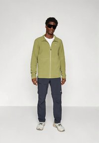 Groene fleece jas met ritssluiting, gecombineerd met marineblauwe cargobroek. Model draagt een zonnebril en beige sneakers. Soepele textuur en een casual pasvorm.