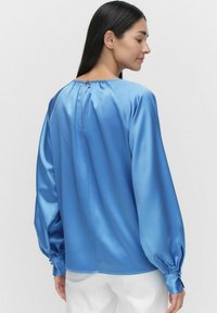 Satinblå blus med långa ärmar, rynkad halsringning, knappstängning bak och åtsittande ärmslut. Slät textur, minimalistisk design.