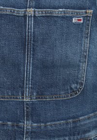 Denimová látka v tmavě modré barvě s viditelnou zadní kapsou a šitými švy. Obsahuje malé vyšití loga s červenými, bílými a modrými akcenty.