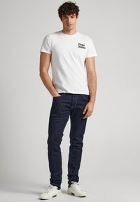 Biely bavlnený tričko s čiernym logom "Pepe Jeans" na hrudi, v kombinácii s tmavomodrými džínsami a bielymi teniskami so zelenými akcentmi.