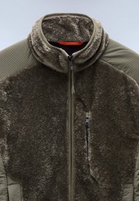 Fleece-takki oliivinvihreänä, jossa on pehmeä tekoturkispinta, korkea kaulus ja teksturoidut sivupaneelit, sekä vetoketjulla varustettu sulkeminen ja taskudetailit.