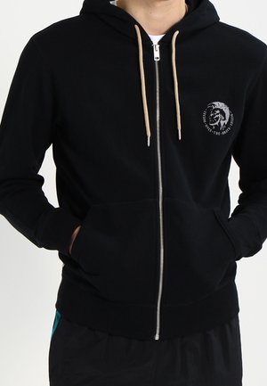Homme portant un sweat à capuche noir zippé avec des cordons beiges et un petit logo sur la poitrine gauche, mains dans les poches avant.