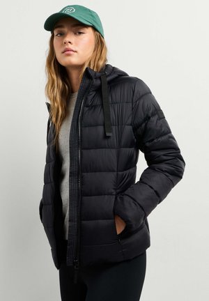 Junge Frau trägt eine schwarze gesteppte Jacke mit Kapuze, graues Shirt, schwarze Hose und grüne Baseballkappe mit Logo, steht vor einem unifarbenen Hintergrund.