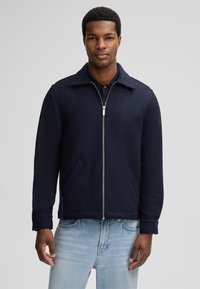 Veste en laine bleu marine avec fermeture éclair, col pointu et deux poches latérales. Présente une coupe structurée et une texture lisse. Portée avec un jean bleu clair.