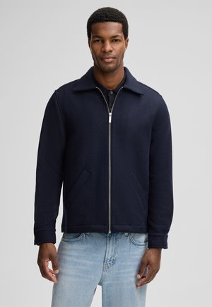 Marineblaue Wolljacke mit Reißverschluss, spitzem Kragen und zwei Seitentaschen. Sie zeichnet sich durch eine strukturierte Passform und glatte Textur aus. Getragen mit hellblauen Jeans.