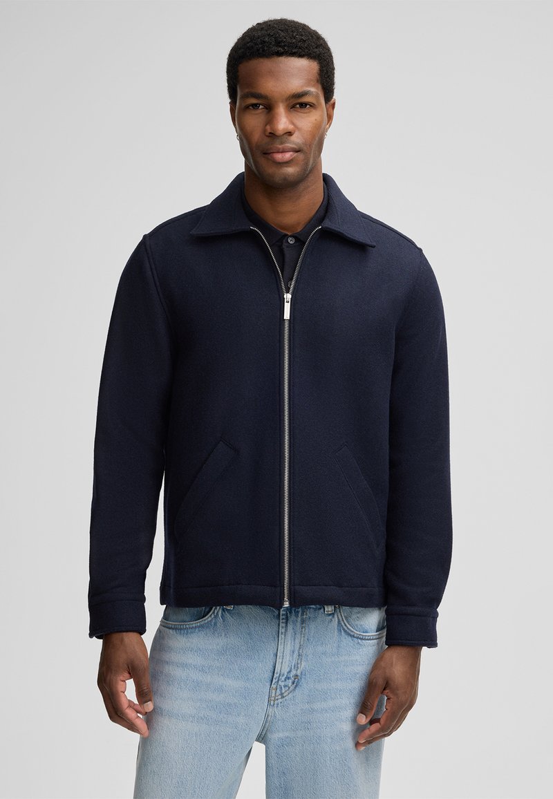 Veste en laine bleu marine avec fermeture éclair, col pointu et deux poches latérales. Présente une coupe structurée et une texture lisse. Portée avec un jean bleu clair.