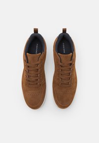 Brown suede sneakers with white rubber soles, featuring a lace-up design, perforated detailing, and navy textile lining.