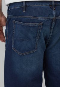Dunkelblaue Denim-Jeans mit einer Gesäßtasche und Steppdetails. Der Stoff hat eine glatte Textur und einen geraden Bein-Schnitt.