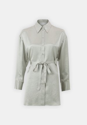 Svijetlozelena bluzica dugih rukava od sjajnog materijala s prednjim kopčanjem na gumbe i vezanom strukom. Ima klasičnu okovratnik i rukave s manžetama.