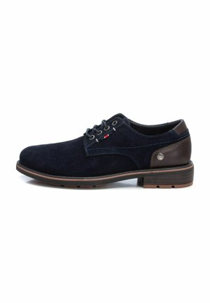 Zapatos de ante azul marino con detalles en cuero marrón, ojales grises y suela a rayas marrones. Presenta costuras azules y un logo decorativo.
