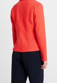 Veste rouge texturée au design ajusté, à manches longues et avec un col. Dotée d'un détail de couture et d'un bouton au niveau du poignet. Assortie à un pantalon sombre.
