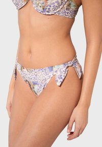 Gros plan sur une femme portant un bas de bikini à motifs léopard violet avec des liens sur les côtés et un haut de bikini assorti sur fond uni.