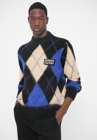 Man in een zwart, blauw en beige argyle trui met een 'Versace Jeans Couture' patch, handen in de zakken, recht vooruit kijkend.