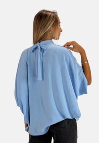 Blusa blu chiaro, strutturata, con una vestibilità ampia e un dettaglio legato sulla parte posteriore. Abbinata a jeans scuri, presenta maniche fluide.