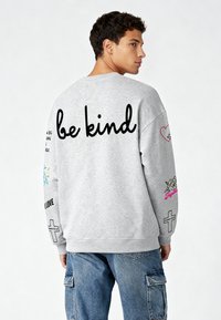 Szürke pulóver laza fitten. A hátsó részen fekete színnel "legyél kedves" felirat, színes grafikákkal és mintákkal a ujjakon.