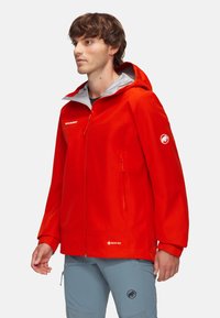 Czerwona kurtka GORE-TEX z kapturem, zamek błyskawiczny z przodu i kieszeniami bocznymi. Posiada szare akcenty oraz logo na rękawie. Gładka, wodoodporna tekstura.