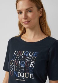 Donkerblauwe t-shirt met korte mouwen, voorzien van de tekst "UNIQUE STORIES" in verschillende kleuren, waaronder goud en blauw, met een ronde halslijn en zachte stof.