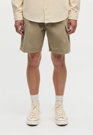 Mężczyzna w beżowych spodenkach typu cargo, kremowej koszuli z długim rękawem i guzikami, ecru skarpetkach oraz kremowych wysokich sportowych butach, stoi na jednolitym tle.