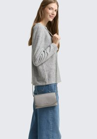 Borsa a tracolla grigia in pelle con finitura testurizzata, forma rettangolare e design slim, tenuta da un modello che indossa una maglia grigia e jeans blu.