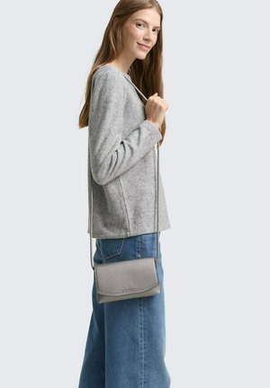 Sac en cuir gris à bandoulière, avec une finition texturée, de forme rectangulaire et au design slim, tenu par un modèle portant un haut gris et un jean bleu.