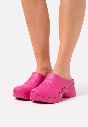 Nohy obuté v jasně růžových klouzavých dřevácích s větranými stranami a vyraženým logem "MK" nahoře, umístěné na bílém pozadí.