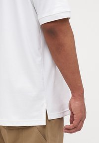 Polo shirt blanc à manches courtes avec fentes latérales, présenté avec un gros plan sur une main sur le côté. Design simple, texture lisse.