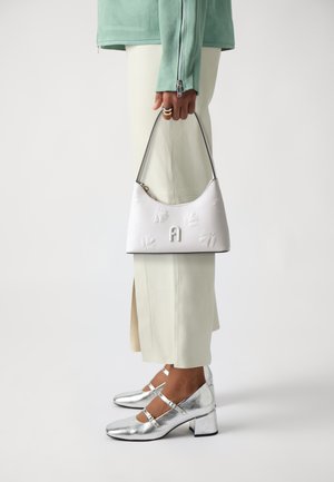 White leather handbag with embossed floral pattern and logo, held by a person in mint jacket and cream pants, paired with silver heeled shoes.