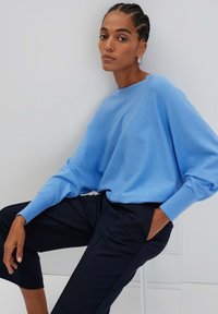 Maglione oversized di colore blu chiaro con colletto alto, maniche lunghe e polsini a costine, abbinato a pantaloni cropped scuri.