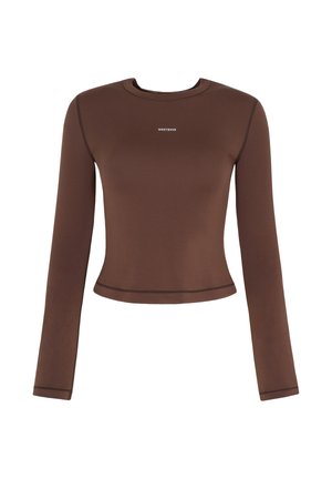 Femme aux cheveux bouclés portant des lunettes de soleil, un crop top marron à manches longues, un pantalon de survêtement gris, des baskets bleues, tenant un sweat-shirt bleu clair sur l'épaule.