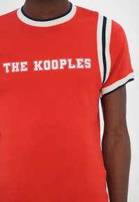 Personne portant un haut rouge à manches courtes avec des bordures blanches et bleu marine, arborant le texte blanc « THE KOOPLES » sur la poitrine.