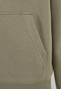 Sudadera color verde oliva con un tejido texturizado, que cuenta con un bolsillo tipo canguro y puños acanalados en el dobladillo. Las costuras son visibles a lo largo de las uniones.