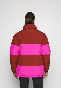 Doudoune à rayures rouges et roses avec un col haut, présentant une texture lisse et soyeuse ainsi que des manches élastiques. Conception pliable avec une coupe décontractée.