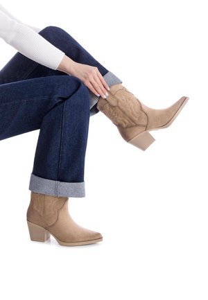 Botas de cowboy de ante color arena con punta afilada y tacón apilado, con detalles bordados. Llevadas con jeans de mezclilla oscura con dobladillo.