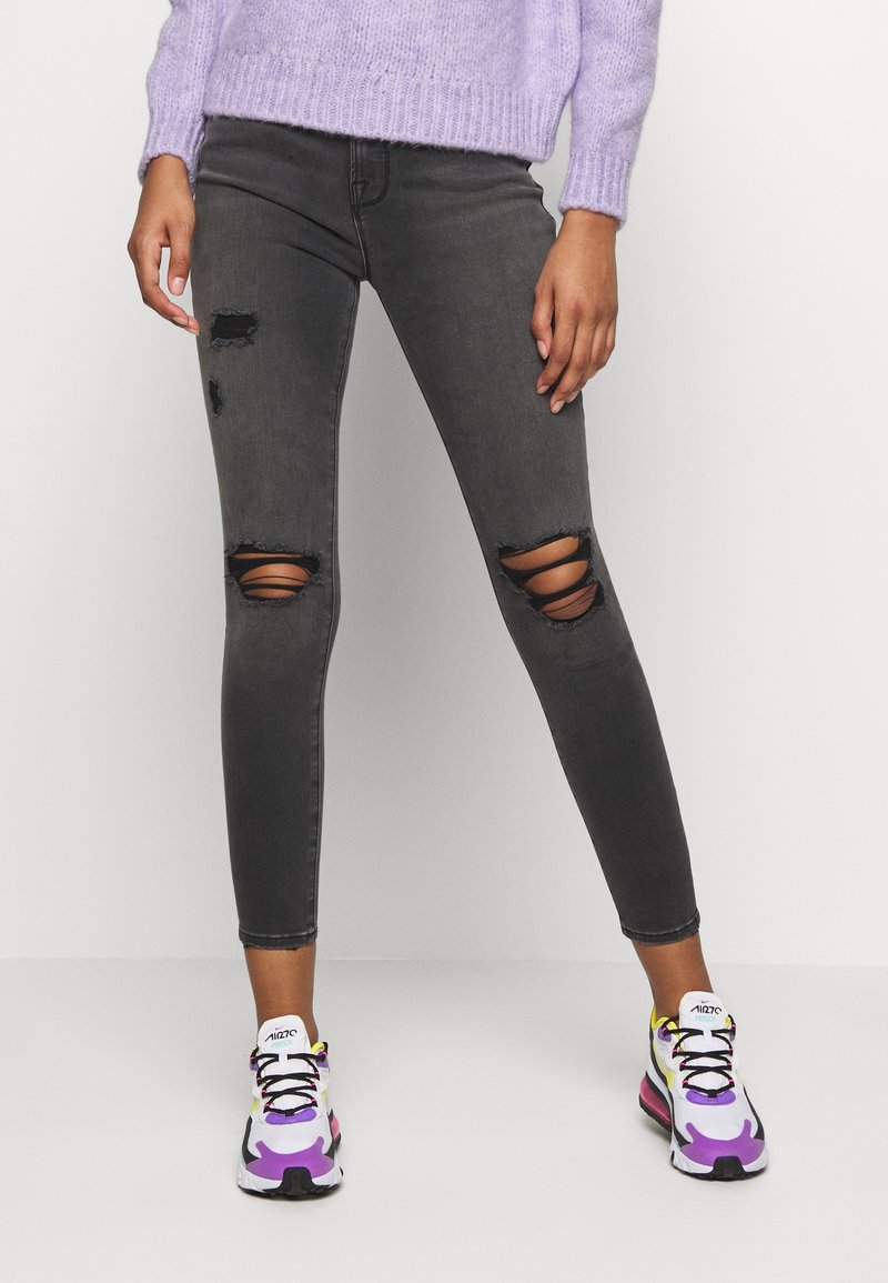 Jean skinny gris avec déchirures aux genoux, associés à des baskets de sport colorées avec des accents violets, jaunes et noirs. Pull en maille texturée.