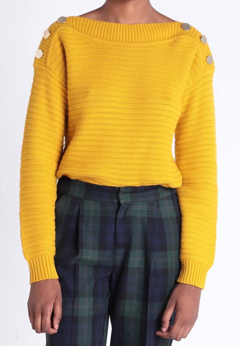 Personne portant un pull côtelé jaune avec des boutons argentés sur les épaules et un pantalon taille haute à carreaux vert foncé et bleu marine.