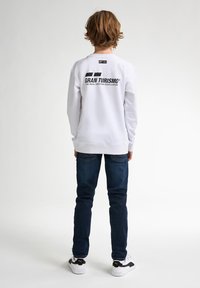 Sudadera blanca con el logo "GRAN TURISMO" en negro en la parte de atrás, combinada con jeans azul oscuro y zapatillas negras y blancas.