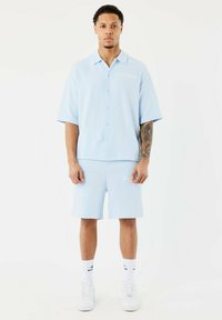Camicia azzurro chiaro a maniche corte con bottoni, caratterizzata da un motivo testurizzato, abbinata a pantaloni corti dello stesso colore. Presenta accenti bianchi del logo e un design minimale.
