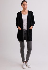 Svart lång cardigan med öppen front, två framfickor och mjukt tyg, i kombination med en vit skjorta och grå skinny jeans.