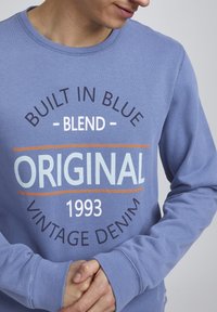 Felpa in cotone blu con scollo rotondo; presenta una grafica audace in bianco, blu navy e arancione: "BUILT IN BLUE," "ORIGINAL," "1993," e "VINTAGE DENIM."
