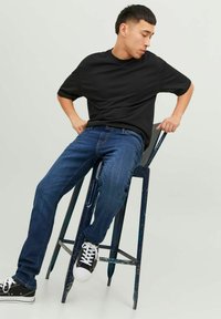 Camiseta negra de manga corta, jeans azules y zapatillas negras. Persona sentada en un taburete de metal azul desgastado contra un fondo neutro.