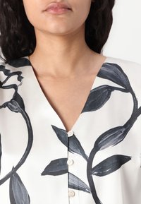 Camicia bianca con scollatura a V, caratterizzata da un design floreale nero e piccoli bottoni. Tessuto liscio con un leggero lucore che enfatizza il motivo.