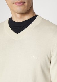 Beżowy sweter z dekoltem w serek wykonany z mieszanki bawełny. Posiada prążkowany dekolt oraz małe haftowane logo „BOSS” na lewej piersi.