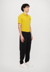 Geel polo-shirt met korte mouwen en een kraag, gecombineerd met zwarte joggers met gestructureerde zijaccenten en witte sneakers.