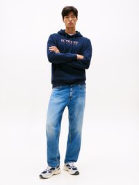 Felpa blu navy con logo "Tommy Jeans", jeans dritti blu chiaro e sneakers bianche/blue, su uno sfondo bianco uniforme.