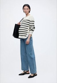 Trudnica koja nosi prugasti džemper sa bočnim gumbima, široke hlače na nozi, crne balerinke i nosi veliku crnu torbu preko ramena.